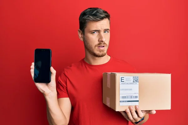 Handsome Caucasian Man Holding Delivery Package Showing Smartphone Screen Shock — Stock Photo, Image