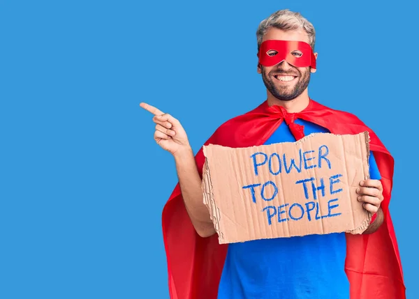 Young blond man wearing super hero custome holding power to the people cardboard banner smiling happy pointing with hand and finger to the side