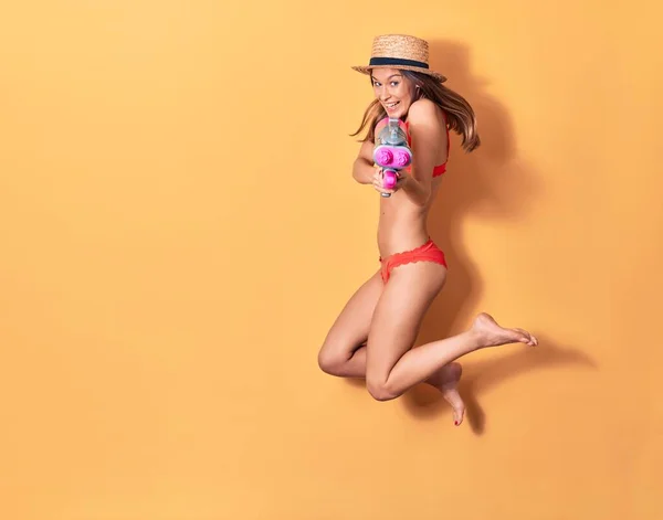 비키니를 모자를 아름다운 소녀가 행복하게 있습니다 배경에 물총을 웃으며 뛰어오르는 — 스톡 사진