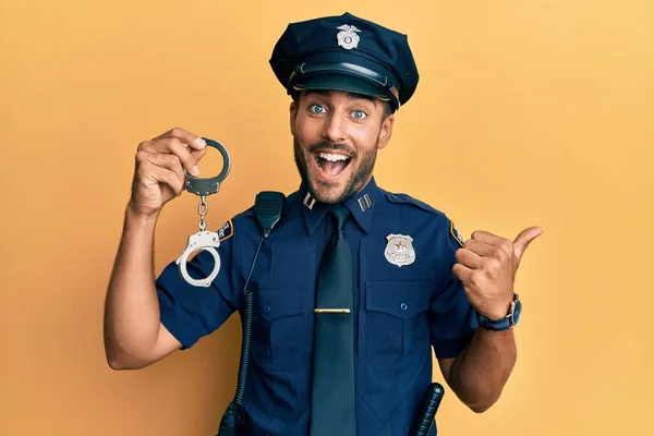Handsome hispanic man wearing police uniform holding metal handcuffs pointing thumb up to the side smiling happy with open mouth