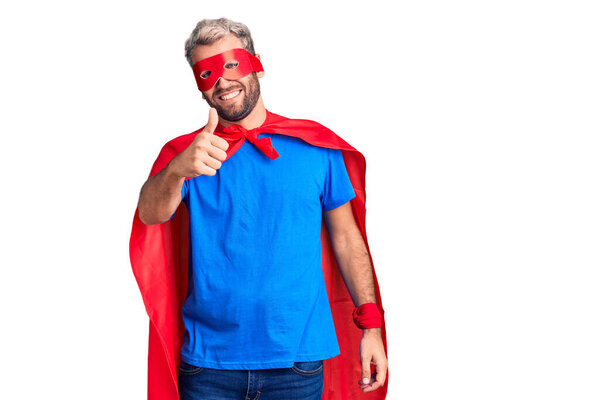 Young blond man wearing super hero custome doing happy thumbs up gesture with hand. approving expression looking at the camera showing success. 