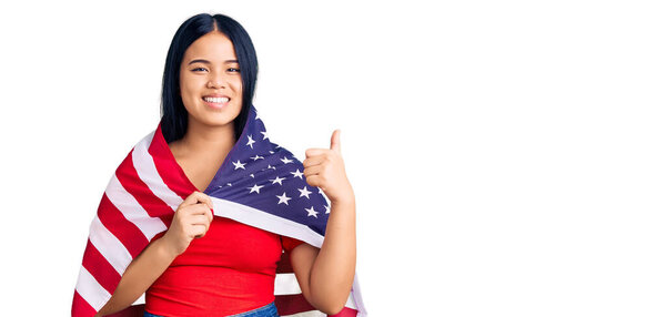 Young beautiful asian girl holding united states flag smiling happy and positive, thumb up doing excellent and approval sign 