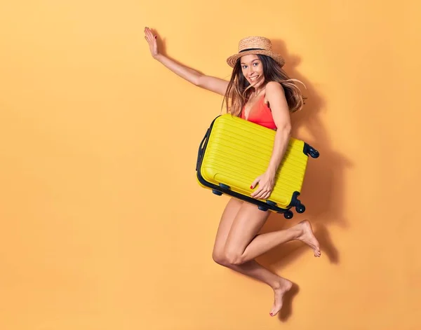 Young beautiful woman on vacation wearing bikini and hat smiling happy. Jumping with smile on face holding cabin bag over isolated background