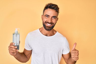 Genç İspanyol adam el dezenfektanı jeli kullanıyor mutlu ve pozitif gülümsüyor, başparmağı mükemmel ve onay işareti yapıyor. 