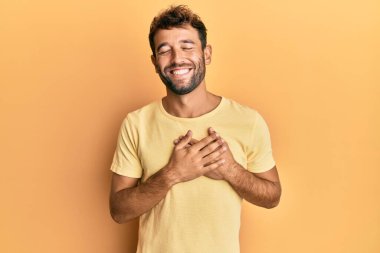 Sarı arka planda sıradan sarı tişörtler giyen, elleri göğsünde, gözleri kapalı ve yüzünde minnet ifadesi olan yakışıklı bir adam. sağlık kavramı. 