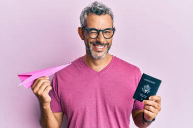 Orta yaşlı, beyaz saçlı, elinde kağıt uçak ve pasaport olan bir adam kameraya seksi bir ifadeyle bakıyor. Neşeli ve mutlu bir yüzü var.. 