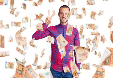 Gündelik kıyafetler giyen genç yakışıklı adam iki numarayı gösterip işaret ederken kendinden emin ve mutlu bir şekilde gülümsüyor..