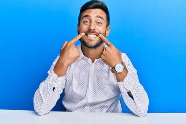 İş elbiseleri giymiş yakışıklı İspanyol bir adam masaya oturmuş ağzı açık gülümsüyor, parmakları işaret ediyor ve neşeli bir gülümseme sergiliyor. 