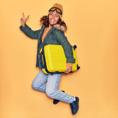 Kışlık giysiler giyen genç, güzel Latin kadın mutlu gülümsüyor. Yüzünde gülümsemeyle kamara çantası tutarak zıplıyor izole edilmiş sarı arka planda zafer işareti yapıyor.