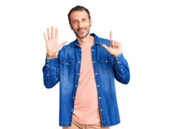 Genç ve yakışıklı bir adam gündelik giysiler giyer ve yedinci parmaklarını göstererek gülümserken kendinden emin ve mutlu olur.. 