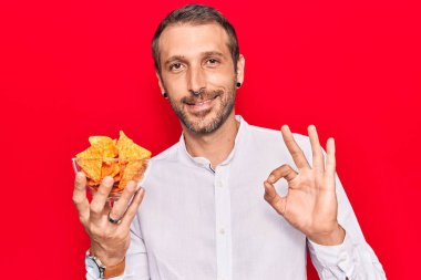 Elinde patates cipsi tutan genç yakışıklı adam parmaklarıyla imza atıyor, gülümseyerek mükemmel bir sembol oluşturuyor. 