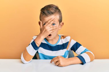 Kafkasyalı sevimli bir çocuk, günlük giysiler içinde masaya oturmuş, şok içinde yüzünü ve gözlerini kapayarak utanmış bir ifadeyle parmaklarının arasından bakıyor.. 