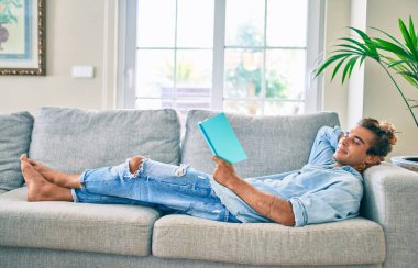 Genç İspanyol gülümseyen mutlu bir kitap okuyucu evdeki kanepede uzanıyor.