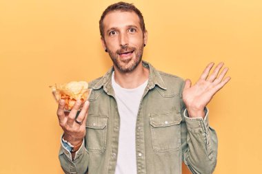 Patates cipsi tutan yakışıklı genç adam başarıyı mutlu bir gülümsemeyle ve kazanan yüz ifadesiyle kutluyor. 