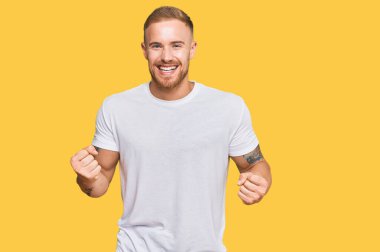 Genç İrlandalı adam gündelik kıyafetler giyip çok mutlu ve heyecanlı bir şekilde kolları havada, gülümseyen ve başarı için çığlık atan kazanan bir jest yapıyor. kutlama konsepti. 