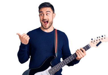 Genç yakışıklı adam elektro gitar çalıyor başparmağıyla yan tarafı işaret ediyor mutlu bir şekilde ağzı açık gülümsüyor. 