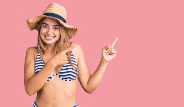 Bikini giyen, şapka takan, gülümseyen ve kameraya bakan genç, güzel sarışın bir kadın.. 