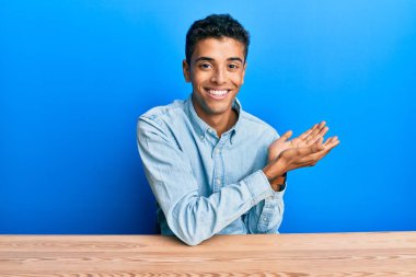 Genç, Afro-Amerikan bir adam, gündelik giysiler içinde masanın üzerine oturmuş avuç içlerini göstererek fotokopi alanını gösteriyor, gülümseyerek mutlu bir şekilde gülümsüyor. 