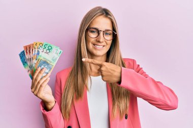 İş stili giyinmiş genç sarışın kadın elinde Avustralya doları tutarak mutlu mesut eli ve parmağı ile işaret ediyor. 