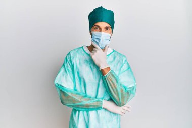 Cerrah üniforması giyen ve tıbbi maske takan genç yakışıklı adam kameraya kollarını kavuşturup gülümseyerek ve çenesini kaldırarak bakıyor. Olumlu düşünmek. 