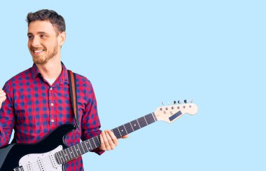 Ayılı yakışıklı genç adam elektro gitar çalıyor baş parmağını yana doğru kaldırıyor ve ağzı açık gülümsüyor. 