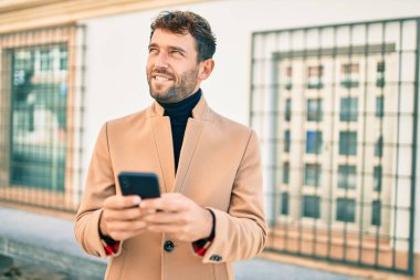 Zarif bir ceket giyen yakışıklı bir iş adamı akıllı telefon kullanarak dışarıda mutlu bir şekilde gülümsüyor.