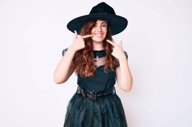 Cadı cadılar bayramı kostümü giyen genç ve güzel bir kadın neşeli bir şekilde gülümseyip parmaklarını ve ağzını işaret ediyor. diş sağlığı kavramı. 