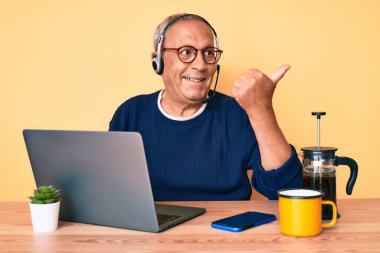 Ofiste çalışan gri saçlı, kıdemli yakışıklı adam operatör kulaklığı takıyor başparmağıyla yan tarafa bakıyor ve ağzı açık gülümsüyor. 