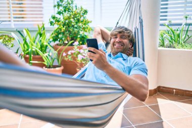 Orta yaşlı yakışıklı bir adam evinin terasında, bir hamakta telefonla dinleniyor.