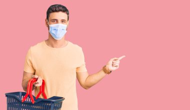 Alış veriş sepeti ve sağlık maskesi takmış yakışıklı genç adam gülümsüyor. 