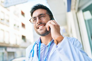 Steteskop takan yakışıklı İspanyol doktor mutlu bir şekilde gülümsüyor. Yüzünde gülümsemeyle kasaba caddesindeki akıllı telefondan konuşuyor..