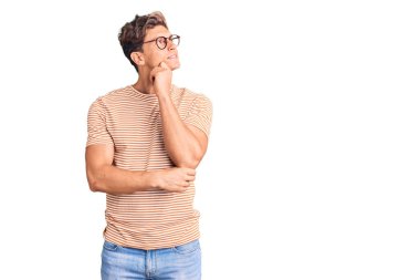 Gündelik kıyafetler giyen ve eli çenesinde gözlük takan genç yakışıklı bir adam soru sormayı düşünüyor, düşünceli bir ifade. Düşünceli bir yüzle gülümsüyor. şüphe kavramı. 