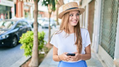 Genç sarışın kadın tatilde gülümsüyor. Şehrin caddesinde akıllı telefon kullanıyor.