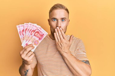 Elinde 100 Yuan 'lık Çin banknotları tutan genç bir beyaz adam ağzını eliyle kapatıyor, şok geçiriyor ve hata yapmaktan korkuyor. şaşırmış ifade 