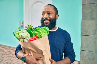 Sakallı Afrikalı Amerikalı bir adam elinde süpermarketten alınmış kağıt poşetler tutuyor.