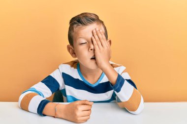 Kafkasyalı sevimli bir çocuk, gündelik giysiler giymiş masada oturuyor. Yarı yüzünü, göz ve ağzını elleriyle kapatarak esniyor. Yüzüm acıyor.. 