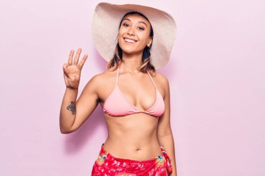 Bikini giyen ve şapkalı genç bir kadın üç numarayı gösterip işaret ederken kendinden emin ve mutlu bir şekilde gülümsüyor.. 