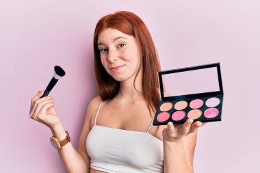 Elinde makyaj fırçası ve allık palet tutan genç kızıl saçlı kız. Hiçbir şeyden habersiz ve kafası karışmış bir ifade. şüphe kavramı. 
