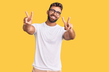 Gündelik kıyafetler giyen ve gözlüklü, ağzı açık gülümseyen genç İspanyol adam zafer işareti yaparken parmaklarını gösteriyor. İki numara.. 