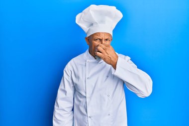 Orta yaşlı, gri saçlı, profesyonel aşçı üniforması giyen ve şapka takan pis ve iğrenç bir koku alan, nefesini burnunda tutan bir adam. Kötü kokuyor. 