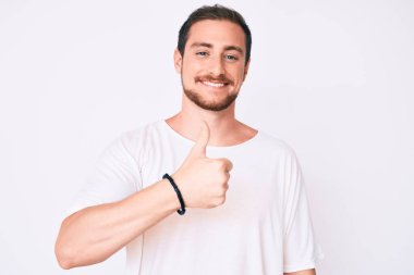 Gündelik beyaz tişört giyen genç yakışıklı adam el hareketleriyle mutlu bir şekilde baş parmak hareketi yapıyor. Kameraya bakıp başarıyı gösteren ifade. 
