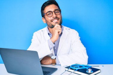 Klinikte çalışan İspanyol üniformalı genç adam kameraya kollarını kavuşturup, çenesini kapatarak gülümsüyor. Olumlu düşünmek. 