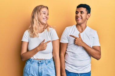 Irklar arası genç bir çift, günlük beyaz tişört giyiyor. Neşeli, yüzünde mutlu ve doğal bir ifadeyle, elleri ve parmaklarıyla yüzünü gösteren bir gülümseme var. 