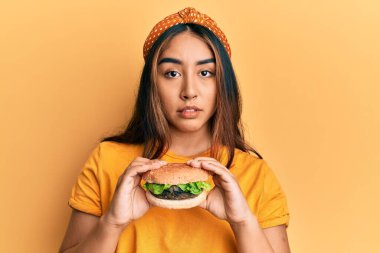 Lezzetli klasik bir burger yiyen genç Latin kadın yüzünde ciddi bir ifadeyle rahatlamış. Basit ve doğal bir şekilde kameraya bakıyorsun.. 