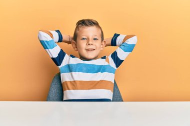 Sevimli beyaz çocuk, günlük giysiler içinde masaya oturmuş, rahatlıyor ve esniyor, kolları ve elleri başının ve boynunun arkasında mutlu bir şekilde gülümsüyor. 