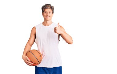 Basketbol topu tutan yakışıklı genç adam mutlu ve pozitif gülümsüyor, başparmağıyla mükemmel ve onay işareti yapıyor. 