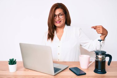 Orta yaşlı esmer, ispanyol bir iş kadını ofiste çalışıyor ve gülümseyen mutlu ve gururlu birini işaret ediyor. 