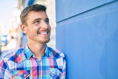 Beyaz bir adam gülümsüyor. Şehrin duvarına yaslanıyor..