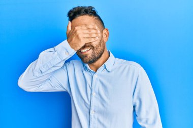 Sakallı, sıradan kıyafetler giyen, gülümseyen ve elleri yüzünde, şaşırtmak için gözlerini kapatan yakışıklı bir adam. kör kavram. 