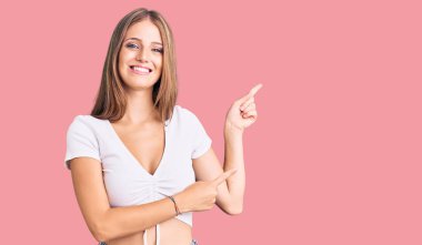 Genç, güzel, sarışın, sıradan, beyaz bir tişört giyen, gülümseyen ve kameraya bakan bir kadın. İki eliyle ve parmaklarıyla kenarı işaret ediyor.. 